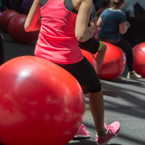 Eine junge Frau trainiert ihre Balance und Rumpfstabilität, während sie auf einem großen roten Gymnastikball sitzt. Was macht man beim Rehasport?