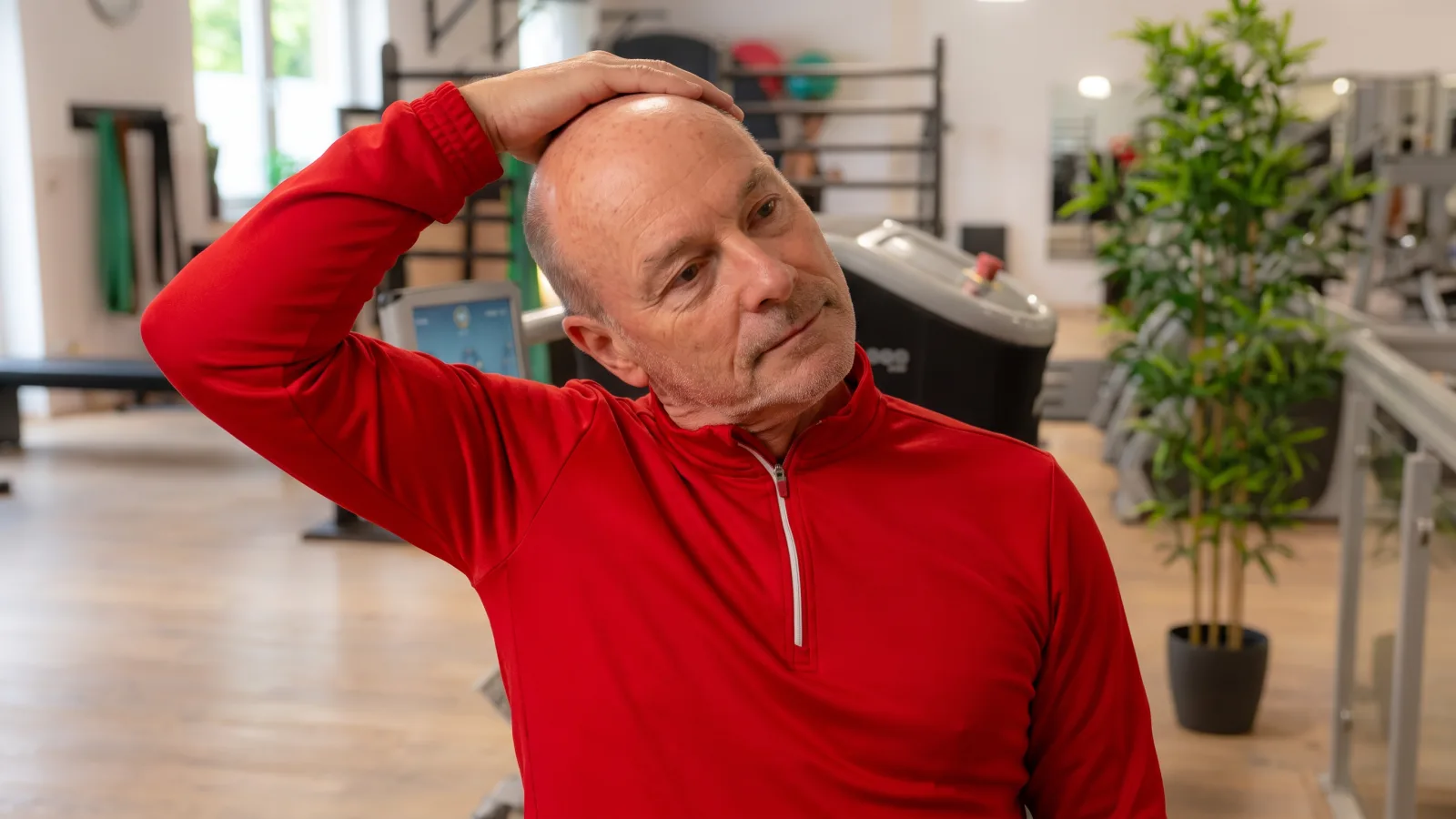 Ein älterer Mann in einem roten Sportoberteil dehnt seinen Nacken in einem Fitnessstudio, um Schmerzen Nacken vorzubeugen oder zu lindern.