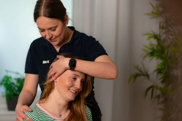 Eine Physiotherapeutin mit dunklem Polo-Shirt behandelt den Nacken einer lächelnden Patientin, die ein grün-weiß gestreiftes Oberteil trägt.