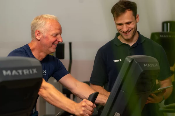 Ein Trainer namens Timon K. lächelt, während er einen älteren Mann beim Training an einem Cardiogerät im Physioaktiv Sportsclub anleitet.